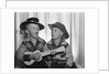 1950s 2 juvenile boys in cowboy hat & shirts playing ukulele & singing mouth open wide by Anonymous