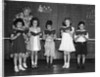 1930s line-up of 5 elementary school students in front of blackboard reading books with teacher looking on by Anonymous