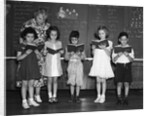 1930s line-up of 5 elementary school students in front of blackboard reading books with teacher looking on by Anonymous
