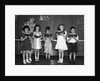 1930s line-up of 5 elementary school students in front of blackboard reading books with teacher looking on by Anonymous