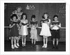 1930s line-up of 5 elementary school students in front of blackboard reading books with teacher looking on by Anonymous