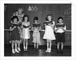 1930s line-up of 5 elementary school students in front of blackboard reading books with teacher looking on by Anonymous
