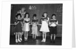 1930s line-up of 5 elementary school students in front of blackboard reading books with teacher looking on by Anonymous