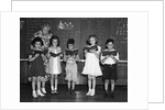1930s line-up of 5 elementary school students in front of blackboard reading books with teacher looking on by Anonymous