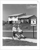 1950s back view of twin girls in plaid skirts & cardigans holding book bags running past school slow sign by Anonymous