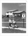 1950s back view of twin girls in plaid skirts & cardigans holding book bags running past school slow sign by Anonymous
