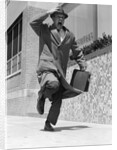 1960s 1950s frantic man running down street holding hat on with hand carrying briefcase wearing top coat by Anonymous