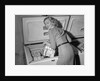 1950s blond woman lifting wire basket food items from a deep freezer looking at camera over her shoulder by Anonymous