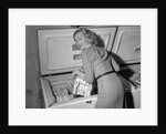 1950s blond woman lifting wire basket food items from a deep freezer looking at camera over her shoulder by Anonymous
