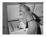 1950s blond woman lifting wire basket food items from a deep freezer looking at camera over her shoulder by Anonymous