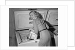 1950s blond woman lifting wire basket food items from a deep freezer looking at camera over her shoulder by Anonymous