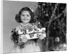 1940s little girl standing near christmas tree holding a wrapped present smiling looking at camera by Anonymous