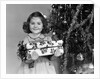 1940s little girl standing near christmas tree holding a wrapped present smiling looking at camera by Anonymous