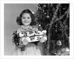 1940s little girl standing near christmas tree holding a wrapped present smiling looking at camera by Anonymous