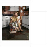 1960s Young Smiling Blond Housewife Sitting On Top Of Laundry Basket Kitchen by Anonymous