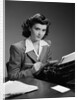 1940s Serious Secretary Stenographer Woman With Pencil Checking Notepad Sitting At Desk With Manual Typewriter Looking At Camera by Anonymous