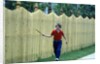 1980s Smiling Boy Running Along Sidewalk Rattling Stick On Tall Picket Fence Making Noise by Anonymous
