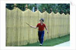 1980s Smiling Boy Running Along Sidewalk Rattling Stick On Tall Picket Fence Making Noise by Anonymous