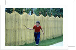 1980s Smiling Boy Running Along Sidewalk Rattling Stick On Tall Picket Fence Making Noise by Anonymous