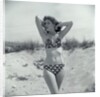 1950s Brunette Bathing Beauty In Polka Dot Bikini Standing In Sand With Hands Behind Head Looking At Camera by Anonymous