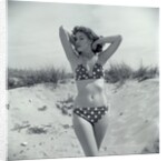 1950s Brunette Bathing Beauty In Polka Dot Bikini Standing In Sand With Hands Behind Head Looking At Camera by Anonymous