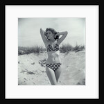 1950s Brunette Bathing Beauty In Polka Dot Bikini Standing In Sand With Hands Behind Head Looking At Camera by Anonymous