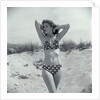 1950s Brunette Bathing Beauty In Polka Dot Bikini Standing In Sand With Hands Behind Head Looking At Camera by Anonymous
