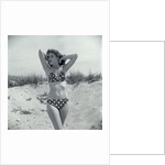 1950s Brunette Bathing Beauty In Polka Dot Bikini Standing In Sand With Hands Behind Head Looking At Camera by Anonymous