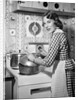 1950s Housewife Wearing Checkered Dress Standing In Kitchen Stirring Pot On Stove Looking Over Shoulder by Anonymous