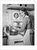 1950s Housewife Wearing Checkered Dress Standing In Kitchen Stirring Pot On Stove Looking Over Shoulder by Anonymous