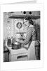 1950s Housewife Wearing Checkered Dress Standing In Kitchen Stirring Pot On Stove Looking Over Shoulder by Anonymous