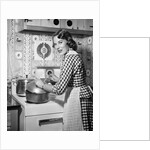 1950s Housewife Wearing Checkered Dress Standing In Kitchen Stirring Pot On Stove Looking Over Shoulder by Anonymous