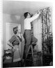 1950s Skeptical Wife In Apron Fingers Pressing Chin Watching Husband On Ladder Hanging New Jungle Motif Drapes In Living Room by Anonymous