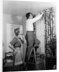 1950s Skeptical Wife In Apron Fingers Pressing Chin Watching Husband On Ladder Hanging New Jungle Motif Drapes In Living Room by Anonymous