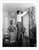1950s Skeptical Wife In Apron Fingers Pressing Chin Watching Husband On Ladder Hanging New Jungle Motif Drapes In Living Room by Anonymous