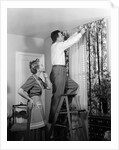 1950s Skeptical Wife In Apron Fingers Pressing Chin Watching Husband On Ladder Hanging New Jungle Motif Drapes In Living Room by Anonymous