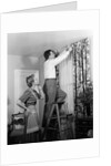1950s Skeptical Wife In Apron Fingers Pressing Chin Watching Husband On Ladder Hanging New Jungle Motif Drapes In Living Room by Anonymous
