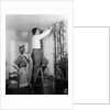 1950s Skeptical Wife In Apron Fingers Pressing Chin Watching Husband On Ladder Hanging New Jungle Motif Drapes In Living Room by Anonymous