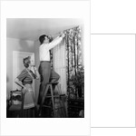 1950s Skeptical Wife In Apron Fingers Pressing Chin Watching Husband On Ladder Hanging New Jungle Motif Drapes In Living Room by Anonymous