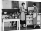 1930s Woman Housewife And Friend Wearing Apron Cooking Food In Kitchen On Gas Stove Indoor by Anonymous