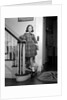 1950s Teenage Girl In Plaid Dress And White Ankle Socks On Stairway Holding Banister Carrying School Books Looking At Camera by Anonymous