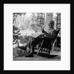 1970s elderly man reading newspaper on porch in a rocker by Anonymous