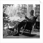 1970s elderly man reading newspaper on porch in a rocker by Anonymous