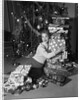 1950s portrait of young woman holding stack of presents next to a Christmas tree by Anonymous