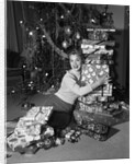1950s portrait of young woman holding stack of presents next to a Christmas tree by Anonymous