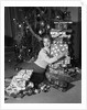 1950s portrait of young woman holding stack of presents next to a Christmas tree by Anonymous
