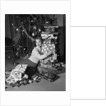 1950s portrait of young woman holding stack of presents next to a Christmas tree by Anonymous