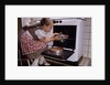 Girl Wearing Apron Removing Cakes from Oven by Anonymous
