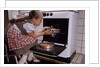 Girl Wearing Apron Removing Cakes from Oven by Anonymous